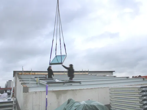 Zwei Männer stehen auf dem Dach eines Rohbaus und nehmen mit den Händen ein Fenster an, das von oben an Ketten befestigt über ihnen schwebt.