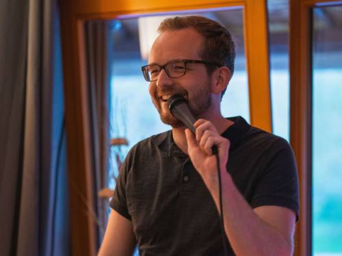 Lachender Mann mit kurzem Haar, Bart und Brille hält ein Mikro.