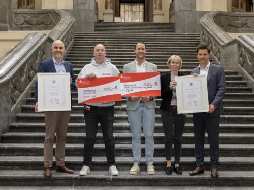 Fünf Personen stehen auf einer Treppe und halten zwei Urkunden und zwei überdimensionale Schecks in den Händen.