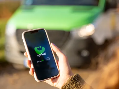 Eine Frau steht im Freien an einer Straße und schaut auf ihr Handy. Ein Transporter nähert sich.