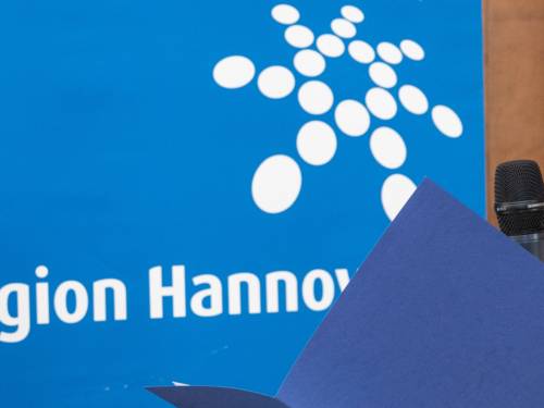 Jemand liest aus einer aufgeschlagenen Mappe und spricht in ein Mikrofon. Im Hintergrund ist das Logo der Region Hannover.