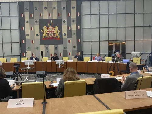 Ratssaal der LHH. Im Vordergrund: 3 Personen sitzen mit dem Rücken zur Kamera. Im Hintergrund: Fünf Personen sitzen der Kamera zugewandt.