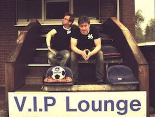 Zwei Männer in Fußballtrikots sitzen auf einer Treppe hinter einem Schild mit der Schrift VIP Lounge.