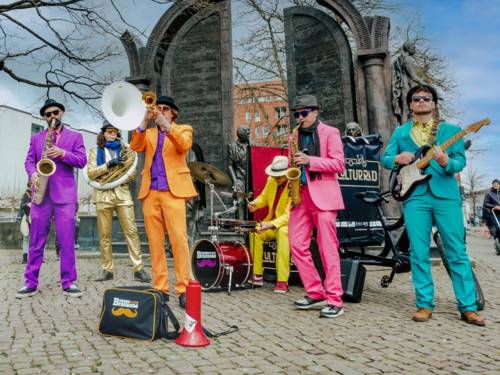 Sechs Musiker tragen pastellfarbene Anzüge und spielen auf einem Platz, vor einem Denkmal, auf ihren Instrumenten.