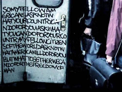 eine mit Text besprühte Tür, Personen mit Taschen in der Hand gehen dadurch.