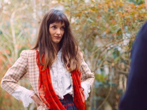 Eine junge Frau in Rüschenbluse, karierter Jacke und gepunktetem Schal schaut - die Hände in die Hüften gestützt - ihr nicht sichtbares Gegenüber herausfordernd an.