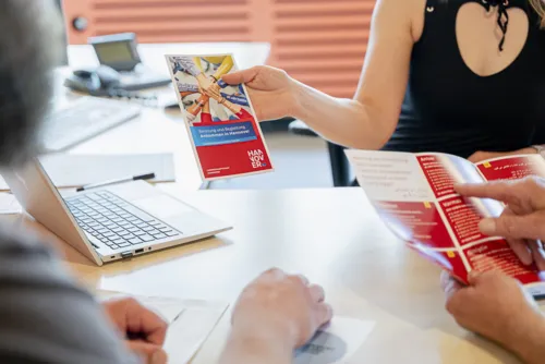 Drei Personen an einem Tisch. Von den Personen sieht man größtenteils nur die Arme und Hände. Auf dem Tisch steht ein Laptop, Die Person hinter dem Tisch hält einen Flyer ins Bild. Die Hände einer weiteren Person auf der rechten Bildseite halten einen weiteren Flyer.