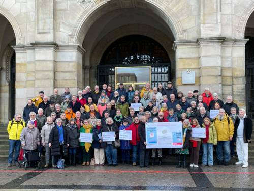 Eine große Gruppe älterer Menschen steht vor und auf einer breiten Treppe vor einem Gebäude mit Bögen über dem Portal, einzele halten Schilder hoch, weitere ein Plakat.