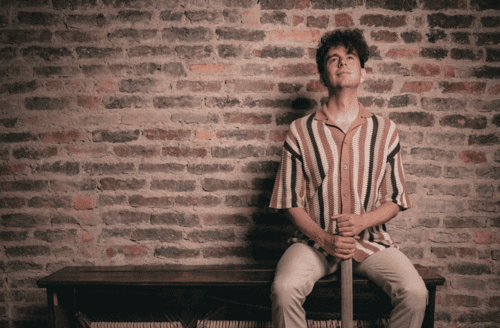 Mann mit kurzen Locken sitzt auf einer Bank vor einer alten Backsteinmauer.