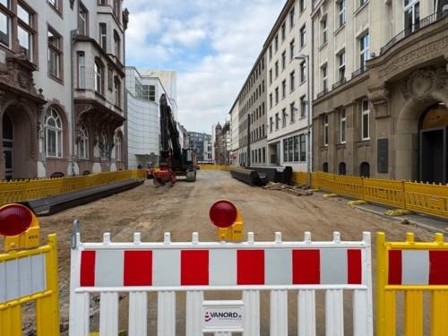 Eine Straße mit abgetragenem Asphalt, vorn eine Baustellenabsperrung, an den Seiten alte schmuckvolle Gebäude.