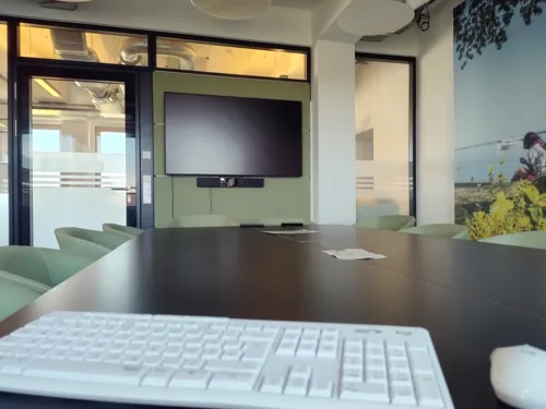 Blick über eine Tastatur und einen großen Besprechungstisch hin zu einem großen Bildschirm, der neben einer Glastür an einer Wand hängt.
