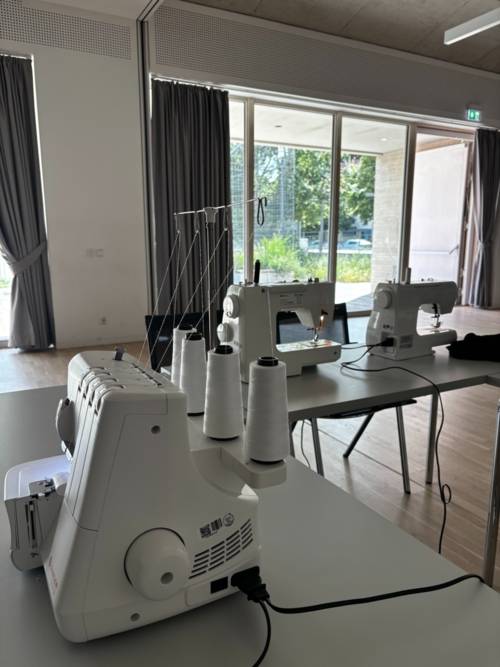 Auf dem Bild sind mehrere Nähmaschinen auf Tischen zu sehen in einem großen Saal.