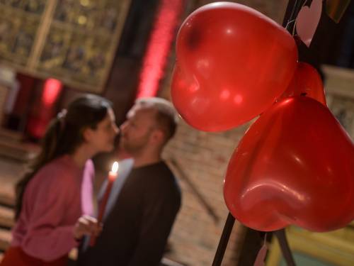 Eine Frau und ein Mann halten in einer Kirche eine Kerze und küssen sich, im Vordergrund hängen Herz-Luftballons.
