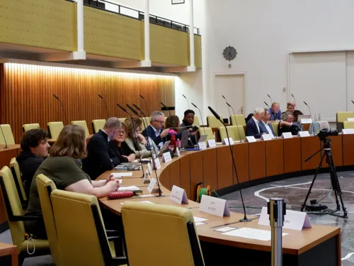 14 Personen sitzen im Halbkreis an den holzvertäfelten Plenumstischen des Ratssaals. Manche unterhalten sich, andere lesen.