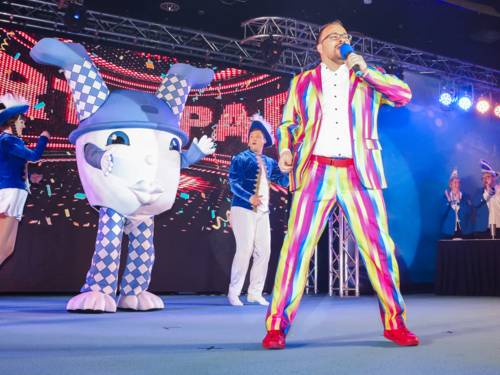 Ein Mann in bunt gestreiften Anzug singt auf einer Bühne in ein Mikrofon, im Hintergrund tanzen eine Person in einem Maskottchen-Kostüm sowie eine Frau und ein Mann in Lindener-Narren-Uniform.