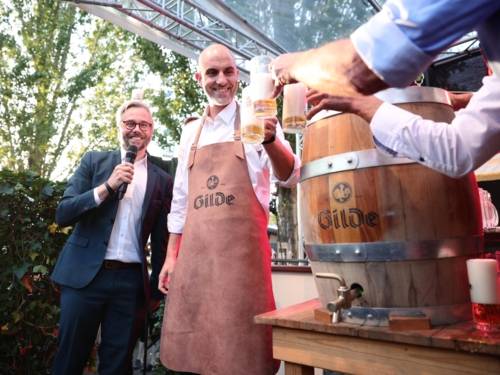 Zwei Männer an einem Bierfass. Am Rand ein dritter, der moderiert. Alle lächeln.