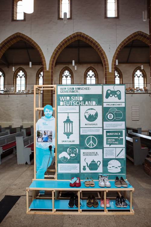Ein improvisiertes Regal in der Mitte eines Kirchenschiffs. An dem Regal mehrere Poster, die hauptsächlich in Türkis gehalten sind. Sie zeigen unter anderem: Ein Gamepad, eine Weltkugel, eine Teekanne, ein Peace-Zeichen. Auf der vorderen Ablage des Regals mehrere Paar Schuhe. Das Poster ganz oben am Regal enthält die Aufschrift "Wir sind Deutschland".