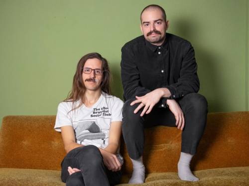 Ein Mann mit langem Haar und Brille sitzt auf einem Sofa, auf der Lehne ein zweiter mit sehr kurzem Resthaar und Schnauzbart.