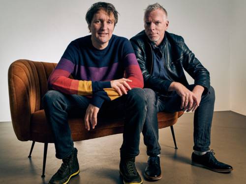 Zwei Männer sitzen auf einem Polstermöbelstück und schauen in die Kamera.