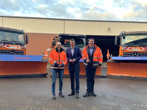 Eine Frau und zwei Männer, dahinter Winterdienst-Fahrzeuge von aha