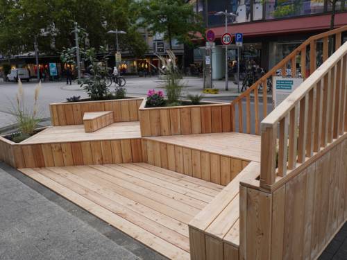 Eine Holzkonstruktion mit Sitzflächen und eingelassenen bepflanzten Blumenkästen an einer Straße.