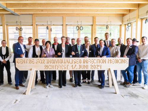 Menschengruppe steht in einem unfertigen Gebäude hinter einem großen Holzbock mit der Aufschrift "Centre for individualised infection medicine"