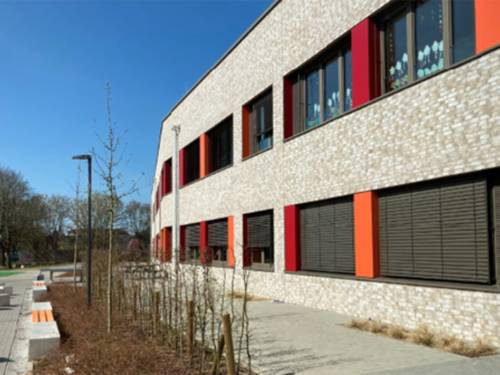 Seitlich ein Flachdach-Gebäde mit hellen Backsteinen und einem Obergeschoss. Die Fensterfronten haben orange und rote Streifen. Links daneben ein Weg, ein Beet mit Sträuchern, davor Bänke.