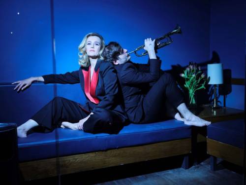 Eine Frau in Anzug sitzt auf einem Sofa. An ihrem Rücken gelehnt sitzt ein Mann, der Trompete spielt.