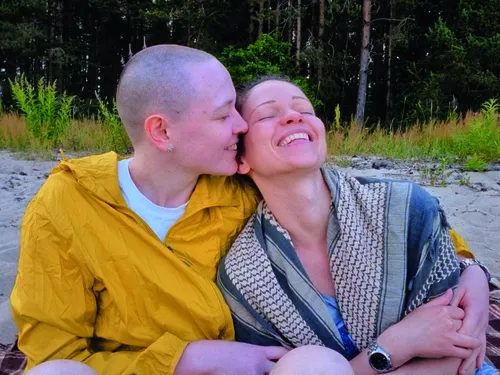 Zwei Personen sitzen gemeinsam an einem Strand vor einer bewaldeten Kulisse. Die nah aufgenommene Szene zeigt eine vertraute Geste, während die leuchtend gelbe Jacke und die natürliche Umgebung farbliche Akzente setzen. Das weiche Licht und die intime Bildkomposition vermitteln eine ruhige, persönliche Atmosphäre.