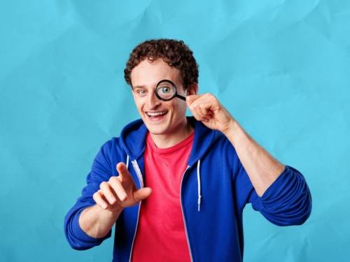 Zu sehen ist Julian Janssen, bekannt als „Checker Julian“. Er trägt eine blaue Hoodie-Jacke und hält eine Lupe vor sein linkes Auge.