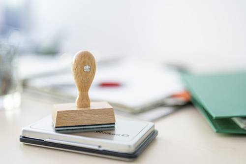 Im Vordergrund im Fokus ein klassische Stempel aus Holz. Er steht auf einem Stempelkissen. Im Hintergrund unscharf Aktenordner.
