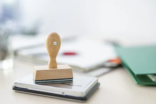 Im Vordergrund im Fokus ein klassische Stempel aus Holz. Er steht auf einem Stempelkissen. Im Hintergrund unscharf Aktenordner.