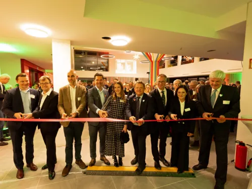 Neun Personen zerschneiden ein rotes Band, im Hintergrund einige hundert weitere Gäste in einem Foyer.