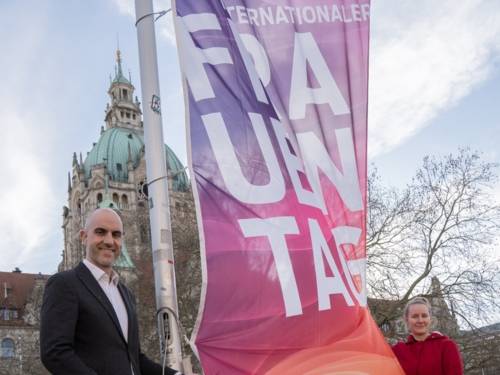 Ein Mann und eine Frau halten eine Flagge in die Kamera. Im Hintergrund ist ein Gebäude zu sehen.