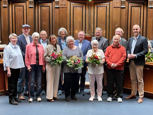In einem Raum mit dunkler Holztäfelung im Hintergrund stehen 13 Personen, einzelne halten Blumensträuße in den Händen.