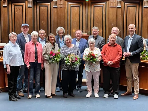 In einem Raum mit dunkler Holztäfelung im Hintergrund stehen 13 Personen, einzelne halten Blumensträuße in den Händen.