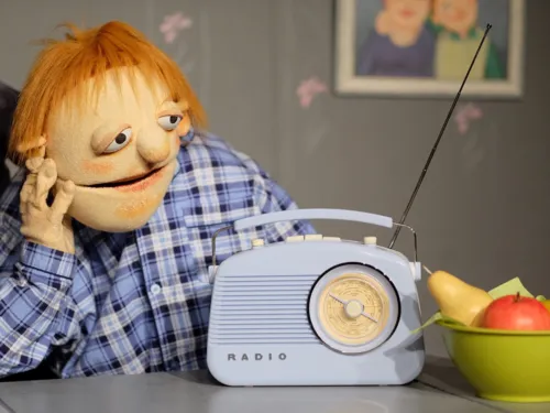 Zu sehen ist eine Handpuppe, vor der ein Radio und eine Schüssel Obst steht.