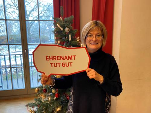 Auf dem Bild ist eine Frau zu sehen, die ein Schild hält mit der Aufschrift „EHRENAMT TUT GUT“. Hinter ihr steht ein geschmückter Weihnachtsbaum.