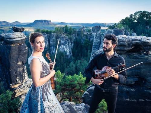 Eine Musikerin und ein Musiker halten jeweils eine Violine und einen Bogen in der Hand, hinter ihnen ist eine Landschaft mit grünen Bäumen, Felsen und einem Abgrund.