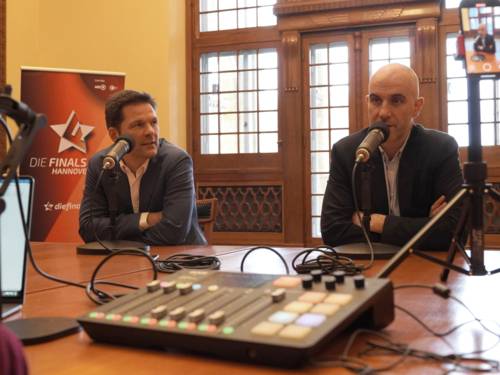 Zwei Männer sitzen an einem Tisch, vor ihnen Mikrofone und ein technisches flaches Bord mit Schaltern und Reglern. Ein Mann spricht, der andere sieht ihn an.