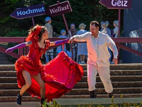 Mann in weißem Anzug und Frau in rotem Kleid auf einer Freilichtbühne, im Hintergrund sind weitere Personen sowie Schilder mit den Aufschriften Hochmut, Wollust und Geiz erkennbar.