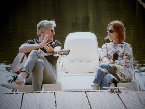 Ein Mann mit Gitarre und eine Frau mit Mikro sitzen in einem Boot an einem Steg.