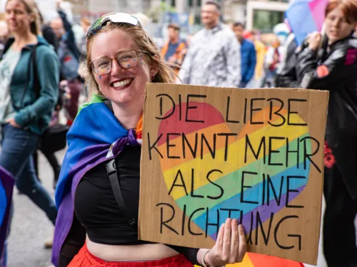 Christopher Street Day in Hannover: Eine teilnehmende Person (Frau) lächelt und hält ein Plakat in die Kamera, auf dem Steht "Liebe kennt mehr als eine Richtung" sowie ein pridefarbendes Herz. Um den Hals hat die Person eine Pride-Flagge gebunden.