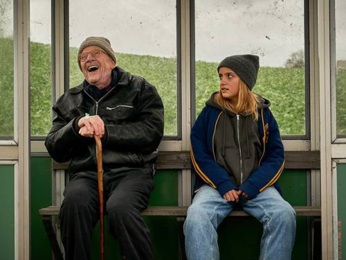 Ein Mann und ein Mädchen sitzen in einem Wartehäuschen. Der Mann lacht schallend, das Mädchen guckt ihn skeptisch an.