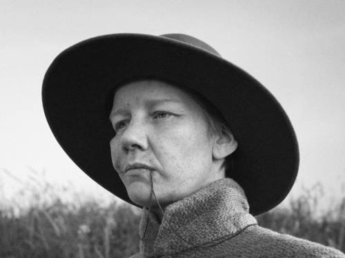 Das Bild zeigt eine Person in einer Schwarz-Weiß-Aufnahme im Freien. Die Person trägt einen breitkrempigen Hut und einen hochgeschlossenen Mantel und ist in Nahaufnahme vor einem unscharfen Hintergrund mit Vegetation zu sehen. Die kontrastreiche Bildgestaltung und die körnige Struktur verleihen der Szene einen dokumentarischen und zeitlosen Charakter.