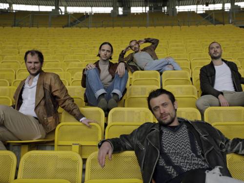 Fünf Männer sitzen auf gelben Sitzen in einem Fußballstadion.