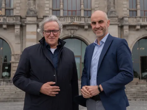 Zwei Personen schauen in die Kamera. Die linke trägt einen Windbreaker und eine Brille, die rechte einen Anzug.