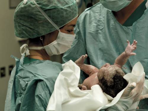 Frau in medizinischer Schutzkleidung h&auml;lt ein Neugeborenes unmittelbar nach der Geburt in den H&auml;nden.