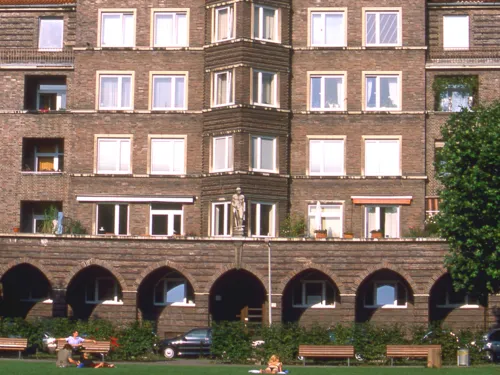 Ein Backsteingeb&auml;ude mit spitz zulaufendem Giebel und Arkaden-Elementen. Davor eine Wiese, auf der sich Menschen aufhalten.