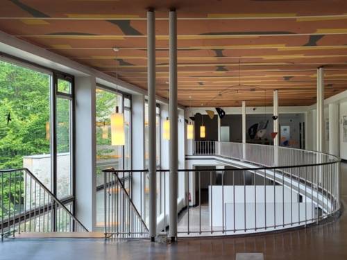 Eine Halle über zwei Geschosse, oben schmale Säulen und ein weißes Metallgeländer mit schwarzem Handlauf, eine Treppe führt hinunter. Links eine große Fensterfront.
